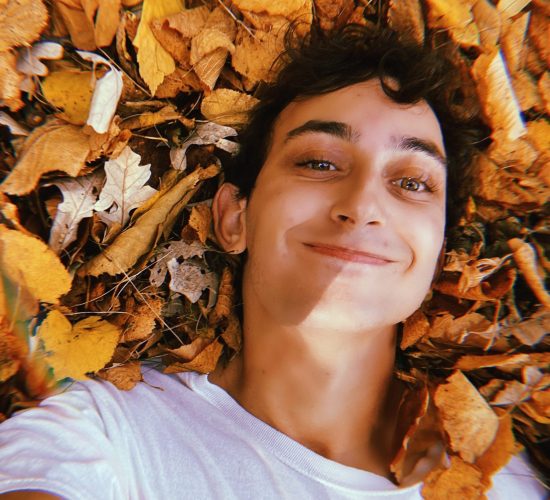 Nicolo smiles for the camera while laying in a pile of yellow and brown leaves. He is wearing a white t-shirt. He has short brown hair.
