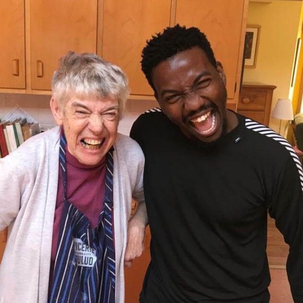 Muka and Mimi stand together with huge smiles on their faces. Mimi is on the left and is wearing a red shirt, a blue apron, and a white cardigan. She has short grey hair. Muka is wearing a black shirt with white stripes down the sleeves. He has short black hair.
