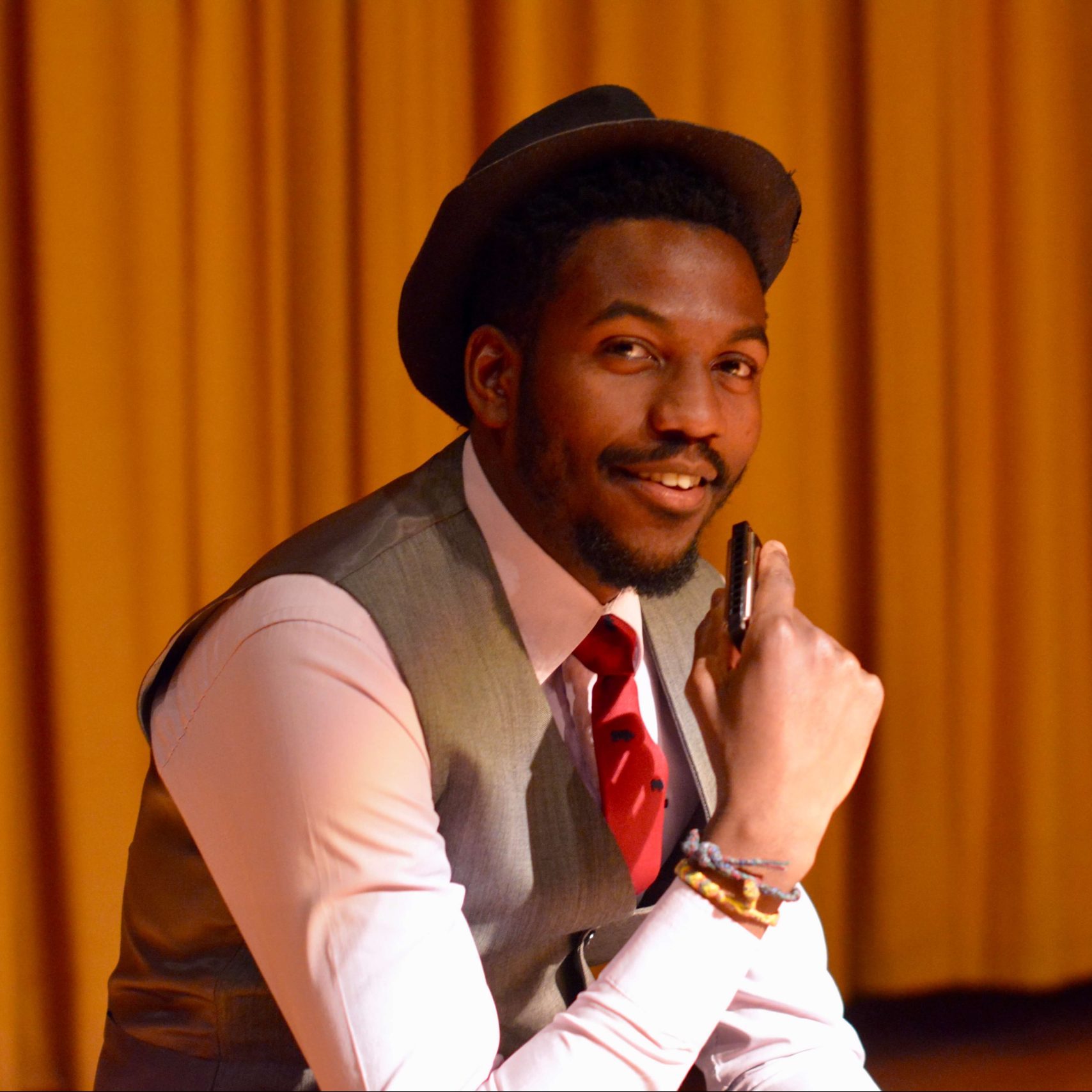 Muka smiles at the camera in a closeup shot while performing in a play. He is wearing a white button up shirt, a red tie, a grey vest, and a brown hat. He has short, dark hair and a beard and moustache. He is holding a harmonica in front of his face, but he is smiling and not playing it.