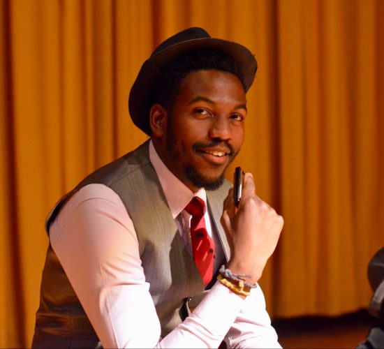 Muka smiles at the camera in a closeup shot while performing in a play. He is wearing a white button up shirt, a red tie, a grey vest, and a brown hat. He has short, dark hair and a beard and moustache. He is holding a harmonica in front of his face, but he is smiling and not playing it.