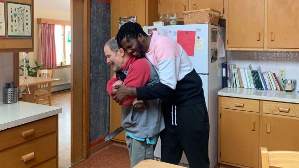 Muka is hugging a friend from behind, and they both have huge smiles on their faces. The friend is wearing a grey and red hoodie as well as grey pants. He has short grey hair and a beard. Muka is wearing black pants and a sweatshirt with black, white, and pink stripes. He has short dark hair and a beard.