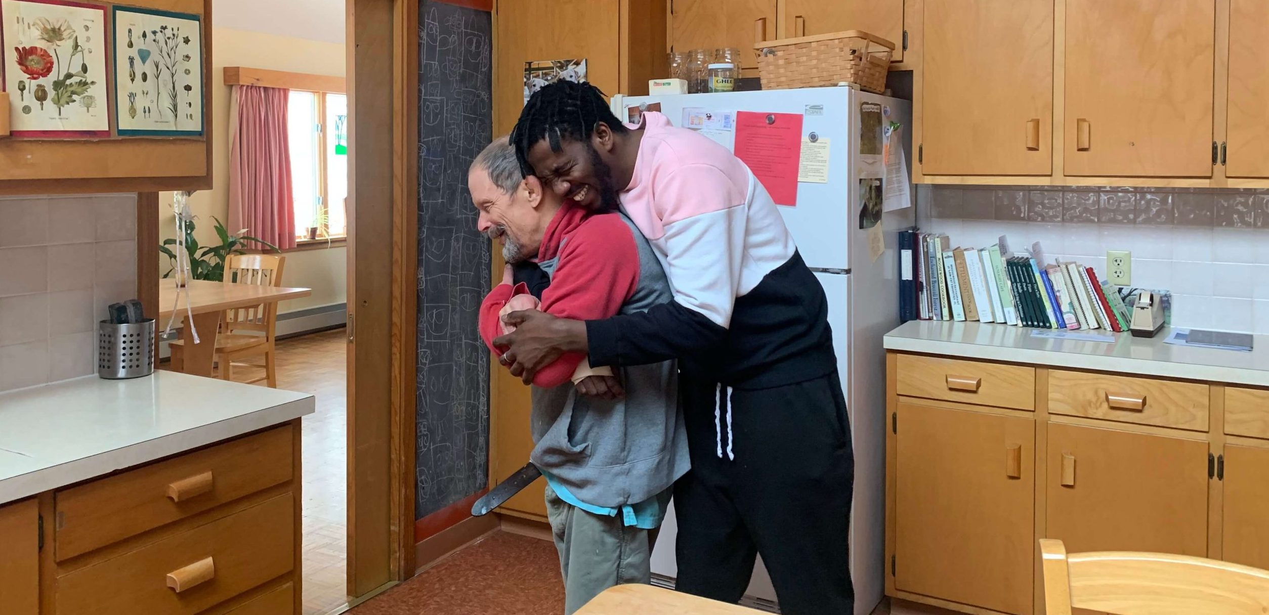 Muka is hugging a friend from behind, and they both have huge smiles on their faces. The friend is wearing a grey and red hoodie as well as grey pants. He has short grey hair and a beard. Muka is wearing black pants and a sweatshirt with black, white, and pink stripes. He has short dark hair and a beard.