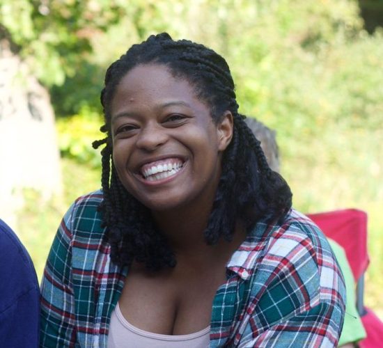 Gabi is smiling for the camera. She is wearing a pink tank top and a green and pink flannel. She is outside, and there is greenery in the background.