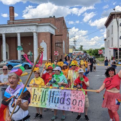 Elias is in the foreground as a crowd of people walk behind him. They are all dressed in colorful clothing and marching in a parade for Gay Pride on Warren street in Hudson New York