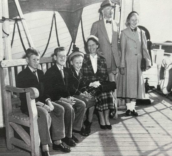 Elias on ship to New Zealand (1955) six people dressed in formal attire pose for a press photograph on a wooden deck. The father and mother are standing on the right side and the children and nanny are sitting on a large wooden bench to the left. This image shows a young Elias and his family aboard a ship