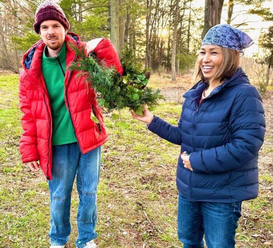 Two people stand in a grassy field with many trees behind them, and they are smiling at the camera. The person on the right is wearing black fuzzy boots, jeans, a blue puffy coat, and a blue patterned bandana. They have light brown hair down to their chin. The person on the left is wearing white sneakers, jeans, a green quarter zip jacket, a red puffy coat, and a maroon and white beanie. They have a short brown beard and are carrying a wreath over their shoulder.