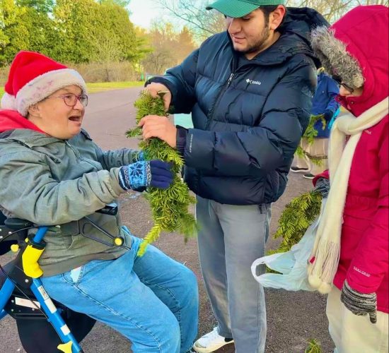 Three people work together to finish making wreaths. On the right are two people standing and looking at a third person, on the left, who is sitting on a blue and yellow walker. They are all wearing heavy coats or jackets, and the person on the left is wearing a red and white Santa hat and blue patterned gloves. The person in the middle and the person on the left are holding a wreath together and making some last placements of branches. The person on the right holds a finished wreaths and listens to the person on the left, who is talking to them.