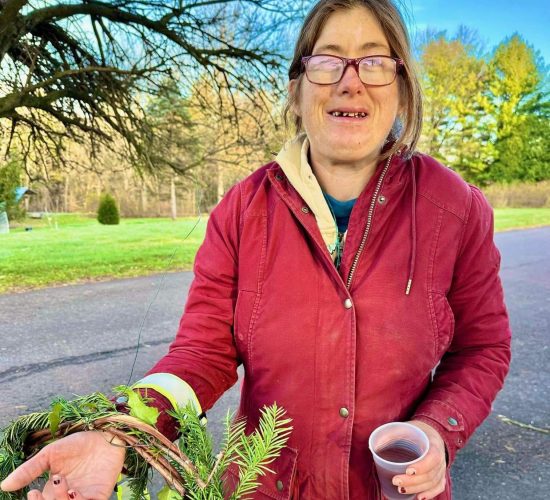 A person smiles at the camera while holding a finished wreath. The wreath has blue, grey, and yellow ribbons tied to it. The person is holding a beverage in their other hand, and they are wearing light blue pants, a red coat, and red glasses. They have brown hair tied into a ponytail behind their head. They are standing on pavement in front of a field of grass and trees.