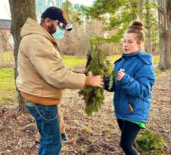 Two people stand in a field of trees and fallen leaves, working together on a wreath. The person on the right is wearing brown fuzzy boots, black pants, and a blue coat with a green shirt poking out from under it, and their light brown hair is tied in a bun on top of their head. The person on the left is wearing brown boots, jeans with a chain on the hip, a light brown coat, a green face mask, and a black and orange hat.