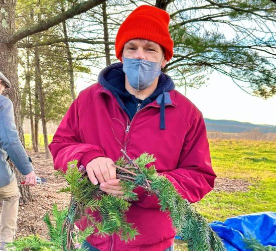 A person smiles at the camera while holding a finished wreath with a green ribbon tied to it in a bow. They are wearing blue pants, a red coat, a black hoodie, a grey face mask, and an orange beanie. They are standing in front of a large pile of branches to be used in wreaths, and there are trees and green fields in the background.