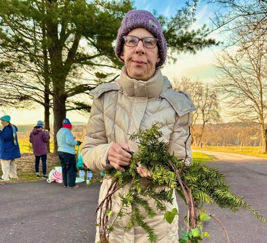 A person smiles at the camera while holding a finished wreath with a red ribbon tied to it. They are wearing white sneakers, jeans, a tan puffy coat, black glasses, and a purple beanie. They are standing on pavement, and in the background are a group of people standing together.