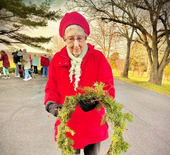 A person smiles at the camera while holding a finished wreath with a red ribbon tied to it. They are wearing black boots, black pants, a red coat, black gloves, a white scarf, and a red beanie. They are standing on pavement, and in the background are a group of people standing together.