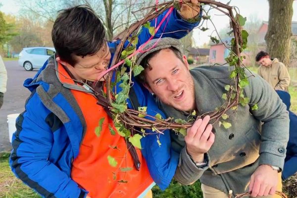 Two people stand in the foreground. One is smiling at the camera and is ducking down to frame their head inside of a wreath. The wreath is being held by the other person, who is smiling down at them and is holding the wreath up above their head.