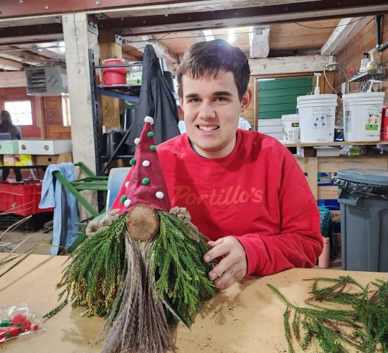 A person sits at a table with a craft in front of them. They are wearing a red sweatshirt that says Portillo's on it, and they are smiling. They have short, straight, brown hair. The craft in front of them is a wooden log with green and brown leaves and branches attached to it. On top of it there is a red felt hat with white and green balls attached to it.