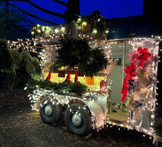 Foxfield Flowers' trailer setup is shown at dusk. The trailer is filled with many wreaths, decorative gnomes, and bows. At the top is a wooden sign that reads "Foxfield Flowers." The trailer is wrapped with white twinkly lights that brighten up the cart and make all of the items and signage visible in the night time. The setup is on gravel ground, and there are silhouettes of tree trunks in the background in front of a dark blue sky.