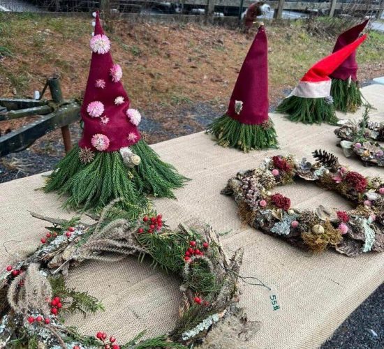 A row of colorful wreaths and felt gnomes sits on a table covered in burlap. The gnomes are made of green branches and have red felt hats. The wreaths have various flowers, berries, and grasses woven into them as decorations. The table is sitting on gravel, and behind its a row of grass, a wooden fence, three parked cars, and a building. The table setup is an extension of Foxfield Flowers' cart at which the items on the table are being sold.