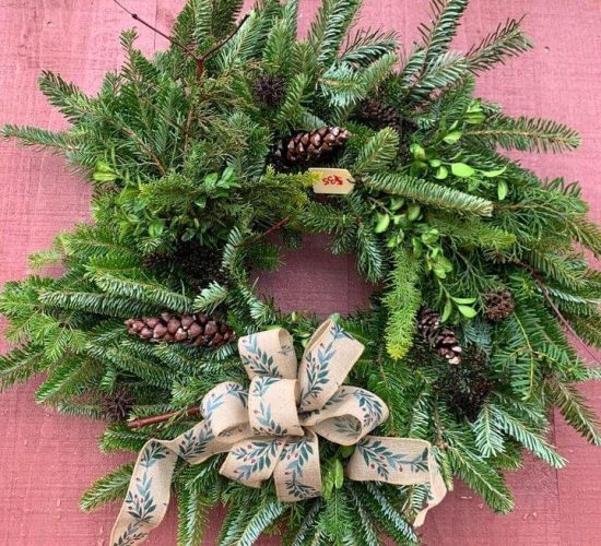 A wreath is pictured hung against a red wall. It is has varied shades of green and varieties of branches in it. There are pinecones scattered throughout the wreath, and at the bottom of the wreath there is a bow of tan ribbon with leaves printed on it. There is a price tag sticking out of the top of the wreath by the center, and it reads $35.