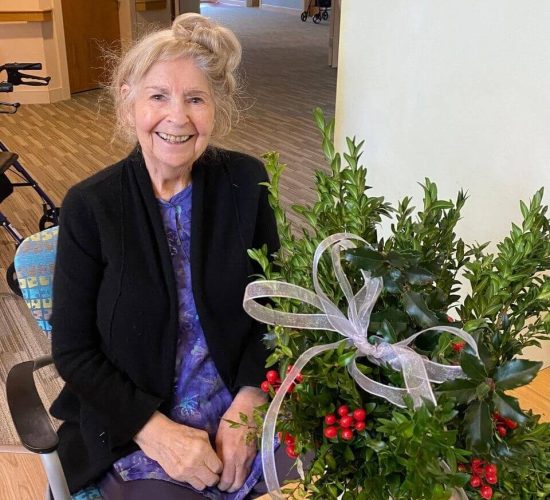 A member of the Camphill Ghent community sits at a table with a boxwood table decoration they've created in front of them. They are smiling at the camera. They are wearing purple pants and a lighter purple shirt with a black cardigan over it. Their long blond hair is on top of their head in a bun. The boxwood table decoration in front of them is filled with branches of green leaves, a few branches of red berries, and a light pink, see-through ribbon tied in a bow.