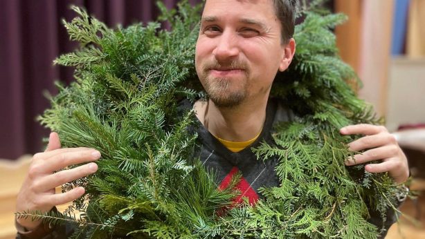 A person smiles and winks at the camera while holding a large green wreath around their neck. They are wearing a red and grey argyle sweater with a yellow tshirt showing underneath it. They have short, straight brown hair and a short beard. They are standing in front of a large purple curtain.