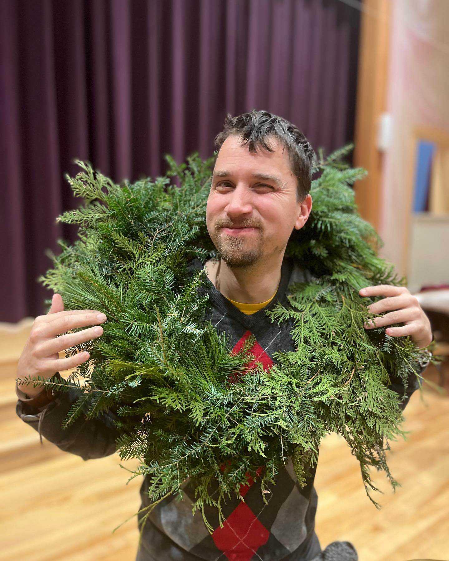 A person smiles and winks at the camera while holding a large green wreath around their neck. They are wearing a red and grey argyle sweater with a yellow tshirt showing underneath it. They have short, straight brown hair and a short beard. They are standing in front of a large purple curtain.