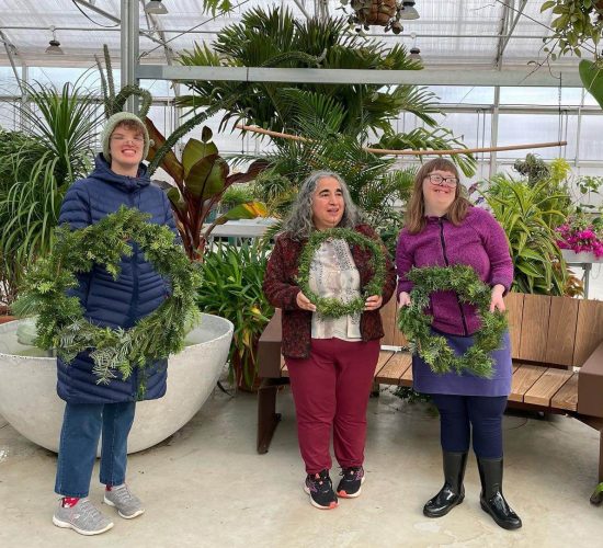 Three people smile at the camera while holding completed wreaths. The person on the left is wearing grey sneakers, red socks, jeans, a blue puffy coat, glasses, and a light green beanie, and they have short, curly, brown hair. the person in the middle is wearing black and pink sneakers, red pants, a white shirt, a maroon jacket, and they have long grey hair. The person on the right is wearing shiny black boots, navy blue leggings, a lavender skirt, a pink jacket, black glasses, and they have long, light brown hair with bangs. They are all standing in front of a large collection of plants and a bench inside of a greenhouse.