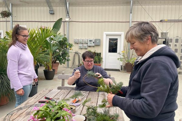 Three people are at a table inside of a greenhouse. The person in the middle is sitting and the two on the sides are standing. The person on the left is wearing lightwash jeans, a light purple turtleneck, black glasses, and their long brown hair in a high ponytail. The person in the middle is wearing a black tshirt, black glasses, and has short brown hair. The person on the right is wearing a black hooded jacket and has short grey hair. They are all smiling, and the person in the middle and right are working on creating wreaths, which they are looking at. Wreath-making supplies are laid out on the wooden table before them, including a wire frame, branches, flowers, and pruning shears.