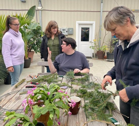 Three people are at a table inside of a greenhouse. The person in the middle is sitting and the two on the sides are standing. The person on the left is wearing lightwash jeans, a light purple turtleneck, black glasses, and their long brown hair in a high ponytail. The person in the middle is wearing a black tshirt, black glasses, and has short brown hair. The person on the right is wearing a black hooded jacket and has short grey hair. A fourth person stands behind the middle person, looking down at them, and they are wearing a green plaid shirt, and a black coat, and they have long brown hair with bangs. The person in the middle and right are working on creating wreaths. The person in the middle is looking up at the person on the left, gesturing to them with their hands while they explain something. Wreath-making supplies are laid out on the wooden table before them, including a wire frame, branches, flowers, and pruning shears.