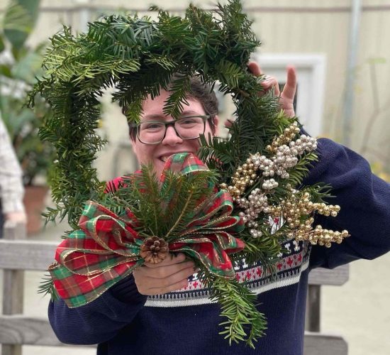 A person smiles at the camera while showing off their completed wreath, which they are holding in front of their face, framing their head. The wreath has a green, red, and gold plaid bow on it with a pinecone in the middle, and gold and silver decorative berries. The person is wearing a blue, grey, red, and black patterned sweater, black glasses, and has short brown hair. The background is out of focus, and they are standing in a greenhouse.